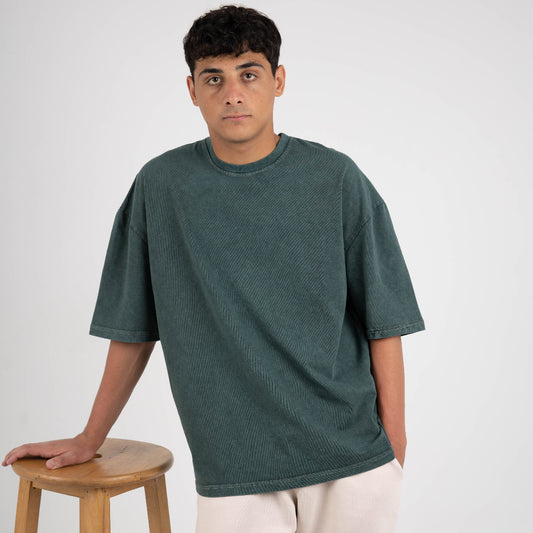 Man wearing a green t-shirt and beige pants standing next to a wooden stool on a white background