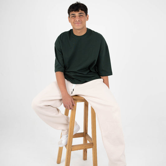 Person sitting on a wooden stool wearing a dark green shirt and light-colored pants against a white background