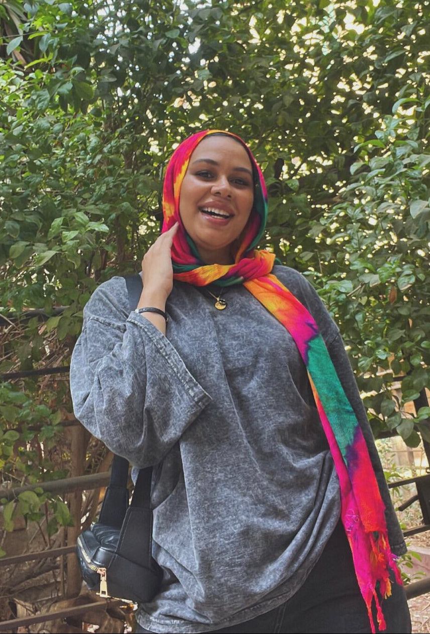 Person wearing a colorful headscarf and gray sweatshirt in front of green foliage